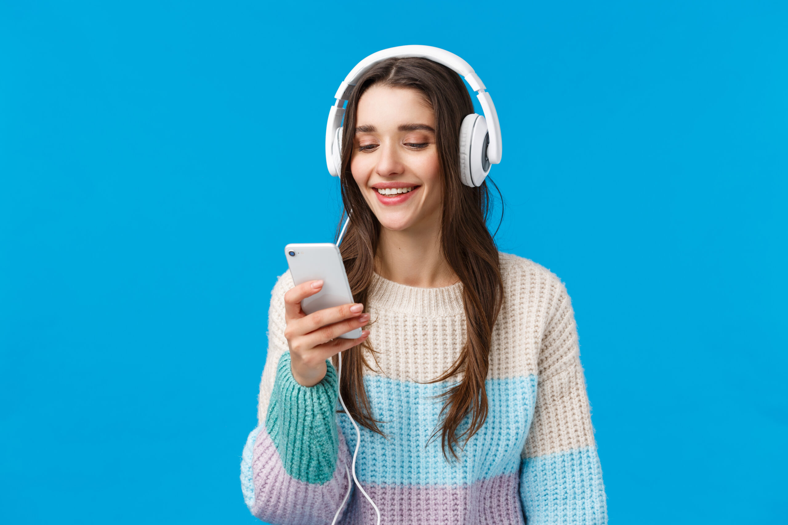 Technology, millennials and lifestyle concept. Carefree cute brunette female student put on headphones, plug-in smartphone picking song and smiling, standing blue background make playlist for study.